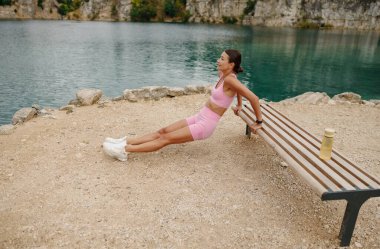 Bir kadın, sakin ve güzel bir göl kenarındaki bankta triseps dalışıyla dışarıda egzersiz yapıyor.