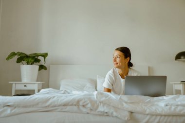 Parlak bir gülümsemesi olan genç bir kadın, davetkar, rahat bir yatak odası ortamında laptopuyla mutlu bir şekilde çalışıyor.