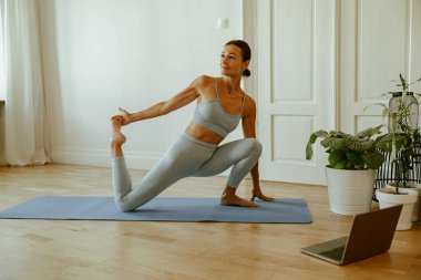 Bir kadın laptopunda yoga kursuyla birlikte evinde yoga pozu veriyor.