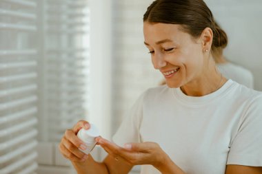 Gülümseyen bir kadın, güzel, modern banyo ortamında ferahlatıcı cilt bakımını yapmaktan mutluluk duyar.