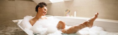 Bir kadın köpük ve suyla dolu bir küvette banyo yaparken gülümsüyor. Banyodaki dinlenme anının tadını çıkarıyor. Musluk ve jakuzinin yanında, eğleniyor.