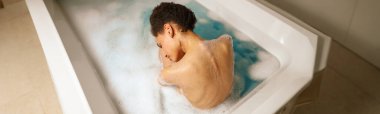 Bir kadın köpüklü suyla dolu bir küvette rahatça banyo yapıyor. Sıvı çıplak göğsünü banyoda sararken dinleniyor.