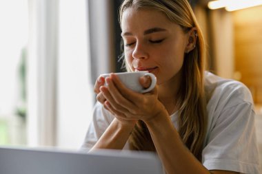 Genç bir kadın sıcak ve davetkar bir atmosferde laptopuyla çalışırken kahvesinin tadını çıkarıyor.