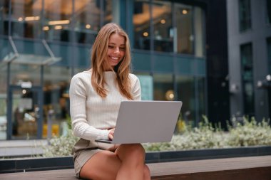 Genç bir kadın, canlı, modern şehir ortamında dizüstü bilgisayarı üzerinde çalışırken gülümseyerek rahatça oturuyor.