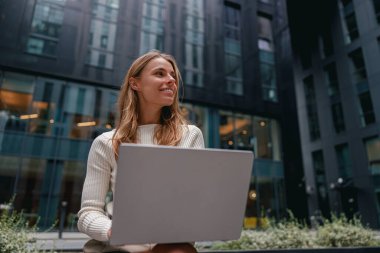 Bir kadın dışarıda oturuyor, mutlu bir şekilde dizüstü bilgisayarını kullanıyor ve gülümsüyor, canlı ve modern bir şehir ortamında.