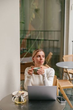 Düşünceli genç bir kadın elinde bir fincan çay tutuyor, sıcak bir kafede işine dalmış.