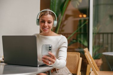 Modaya uygun genç bir kadın, rahat bir kafede vakit geçirirken bilgisayarını ve akıllı telefonunu aktif olarak kullanıyor.