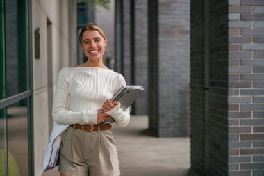 Çağdaş bir ofis ortamında duran neşeli, genç bir kadın elinde bir defter tutuyor.