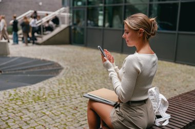 Modaya uygun bir kadın bir bankta oturuyor, akıllı telefonuyla meşgul, modern mimari ile çevrili..