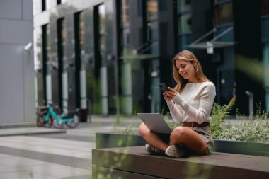 Genç bir kadın laptopuyla meşgul olurken aynı zamanda telefonunu açık havada canlı bir şekilde kullanıyor.