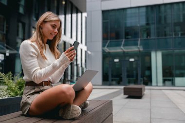 Genç bir kadın, şehir dışındaki bir çalışma alanında bulunduğu sırada aktif olarak dizüstü bilgisayarıyla ve akıllı telefonuyla meşgul.
