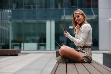 Genç bir kadın dışarıda dinlenirken hem akıllı telefon hem de dizüstü bilgisayar kullanıyor.