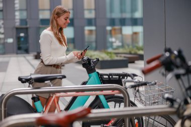 Genç bir kadın, canlı, modern bir şehir manzarasında bisikletin yanında duruyor ve akıllı telefonunda güncellemeleri kontrol ediyor.