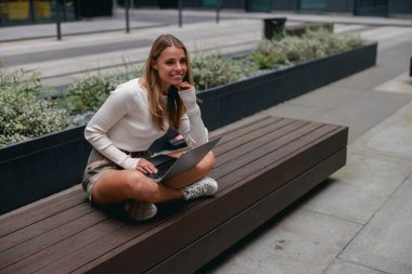 Genç bir kadın, tamamen şehir merkezine odaklanmış, modern bir ortamda otururken bilgisayarına dalmış durumda.