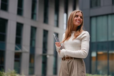 Elinde dizüstü bilgisayarla gülümseyen şık bir genç kadın, çağdaş bir şehir ortamında zarif bir şekilde giyinmiş..
