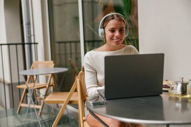 Şık kulaklıklar takan genç bir kadın, modaya uygun, modern bir kafede laptopuyla çalışmaktan zevk alıyor.