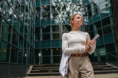 Kendine güvenen bir kadın elinde defteriyle modern cam ofis binasının önünde gururla durur.