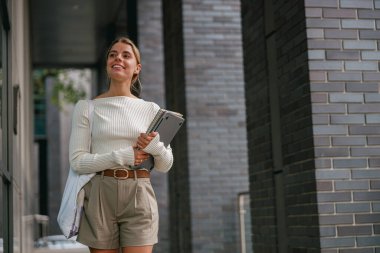 Kendine güvenen ve stil sahibi genç bir kadın bir defter taşıyor. Canlı ve modern bir şehir ortamında yürüyor.