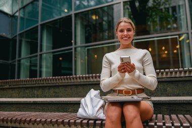 Gülümseyen bir kadın akıllı telefonuyla, canlı bir şehir yaşam tarzı ve gelişmiş teknoloji sergiliyor.