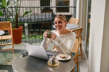 Genç bir kadın şık bir kafede dizüstü bilgisayarla çalışırken kahvesinden zevk alıyor.