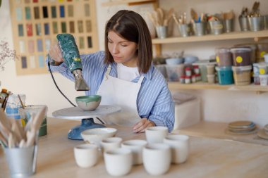 Bir çömlekçi, canlı bir sanat stüdyosunda seramik bir eser yaratıyor. Etrafında aletler ve malzemeler var..