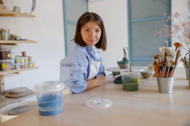 Bir sanatçı işine derinden odaklanmış, çeşitli araç gereçler ve malzemelerle dolu canlı bir stüdyoda boya uyguluyor.
