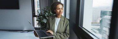 Bir beyazlar işçisi bir dizüstü bilgisayarla pencerenin yanında duruyor ve onu bir çıkış aracı olarak kullanıyor. Onun yanındaki saksıda bir saksı var.