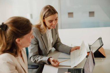 Gösterişli ve modern bir ofis ortamında, iki kadın proje belgeleri ve fikirleri etraflıca tartışırken görülebilir.