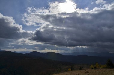 Sonbahar dağ manzarası, ormanın arkasındaki tepelerin karanlık siluetleriyle bulutlu gökyüzünün altında güneş ışınlarıyla. Karpat Dağları, Ukrayna