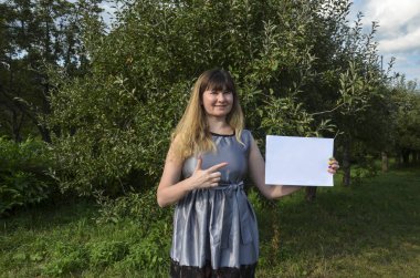 Gülümseyen, şık, esmer bir kızın elindeki beyaz kağıt parçasına bakıp doğal arka planda poz vermesi ön planda. Reklam kavramı.