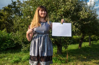 Gülümseyen, şık, esmer bir kızın elindeki beyaz kağıt parçasına bakıp doğal arka planda poz vermesi ön planda. Reklam kavramı.