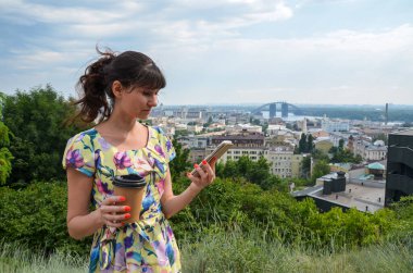 Çekici, gülümseyen, cep telefonuyla kahve içen ve parktaki kameraya bakan genç bir kadın.