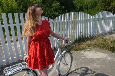 Smiling young and beautiful long-haired girl in red dress standing with retro bike at the summer garden