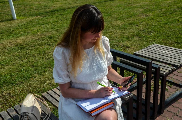 Elragadó Csinos Lány Egy Padon Parkban Belenéz Telefonba Egy Jegyzetfüzetbe — Stock Fotó