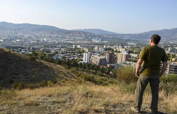 Back view of man standing on the hill and looking to the residential buildings and mountains in background at Tbilisi, Georgia