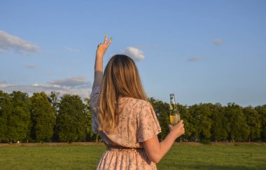 Gün batımında yeşil çimlerin üzerinde duran bir şişe birayla arkadaşlarını bekleyen bir kızın arka görüntüsü. Tatil ve dinlenme