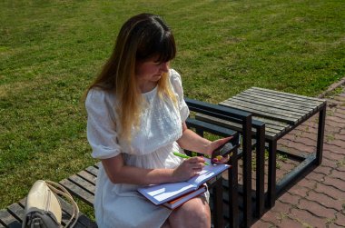 Not defterleri ve kitapları olan çekici bir kız bir bankta oturur ve telefona bakar. 