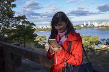 Akıllı telefon kullanan güzel bir kadın, telefonda kısa mesajlar okuyor ve şehir doğasına karşı parkta arkadaşlarıyla sohbet ediyor. Modern teknoloji kavramı