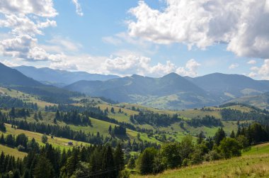 Bir vadideki küçük bir yerleşim yerinin etrafındaki yeşil orman. Bulutların arasından geçen bir güneş ışını dağların ve vadinin farklı bölgelerinde bir ışık ve gölge oyunu yaratıyor. Karpatlar