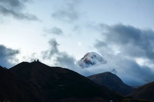 View from Stepantsminda to silhouette of the Gergeti Monastery against the backdrop of majestic mount Kazbek with snow-capped peak, veiled in misty clouds. Travel to Georgia
