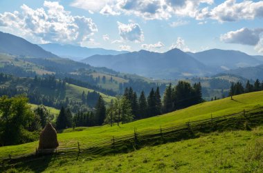 Yuvarlanan yeşil tepeler, geleneksel ahşap bir çit ve görkemli dağlardan oluşan bir saman yığını ve kabarık bulutlarla kaplı bir gökyüzü içeren resim manzarası. Karpatlar, Ukrayna
