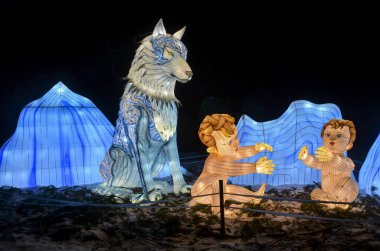 Buzlu dağların arkasında bir kurdun yanında oynayan iki çocuk figürleriyle dolu gece manzarası. Işık heykelleri ve kurulumları festivali