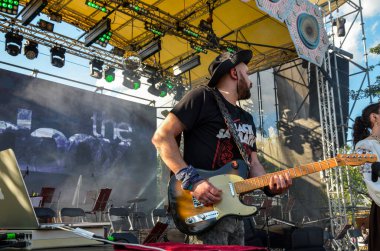 Dinamik canlı müzik sahnesinde ön planda bir gitarist, etrafında müzik ekipmanları olan bir elektro gitar çalıyor, canlı bir olayın arka planında geçiyor. 