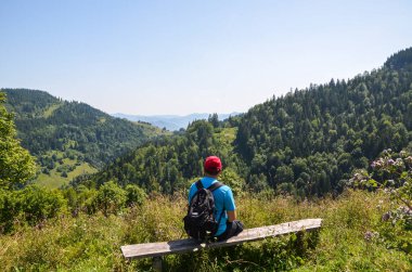 Sırt çantalı bir adam, ahşap bir bankta oturuyor, kameradan uzağa bakıyor, yuvarlanan tepeleri, dağları ve açık mavi gökyüzünün altında yoğun ormanları olan yemyeşil bir vadiye bakıyor. 