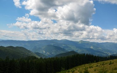 Ön planda çimenli bir tepenin yer aldığı görkemli manzara, parçalı bulutlu bir gökyüzünün altında yoğun ormanlık dağlara yol açıyor. Karpatlar, Ukrayna