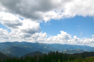 Ön planda çimenli bir tepenin yer aldığı görkemli manzara, parçalı bulutlu bir gökyüzünün altında yoğun ormanlık dağlara yol açıyor. Karpatlar, Ukrayna
