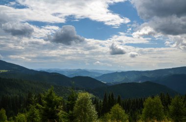 Önde yeşil ağaçlar ve arka planda çok katmanlı dağ sırtları olan sık ormanlarla kaplı yuvarlanan tepeler yer almaktadır. Ukrayna Karpatlarının dağlık bir bölgesinin nefes kesici manzarası