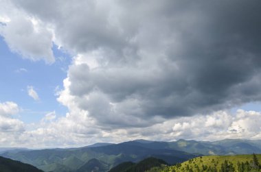 Yukarıdaki görkemli gökyüzü altında sık ormanları barındıran manzara, kabarık beyaz bulutlar ve daha koyu, daha yoğun bulutlarla dolu. Bu da yaklaşan bir fırtına veya yağmur olduğunu gösteriyor.
