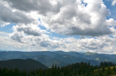 Kümülüs bulutlarıyla dolu dinamik bir gökyüzünün altında sık ormanlarla kaplı dağ sırtı manzarası. Işığın ve gölgenin etkileşimi derinlik katıyor ve doğal alanın enginliğini vurguluyor.