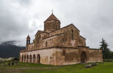 Historic Odzun church stands under a cloudy sky, surrounded by lush green grass and distant mountains, evoking a sense of serenity and timeless beauty. Lori Region, Armenia clipart